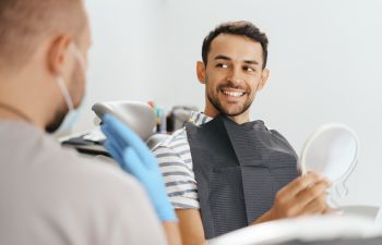 A satisfied mman in a dental chair looking at his restored smile in a mirror and a dentist giving post-treatment instructions.