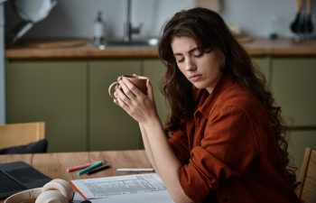 A woman sits at a table, holding a cup while contemplating her thoughts in a cozy kitchen.