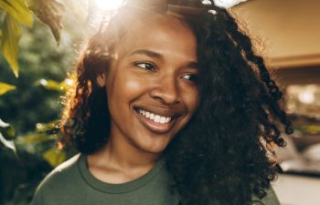 Facial close-up portrait of youthful African female walking down streets of favorite neighborhood, posing against green trees, smiling widely, feeling free, enjoying hot summer day
